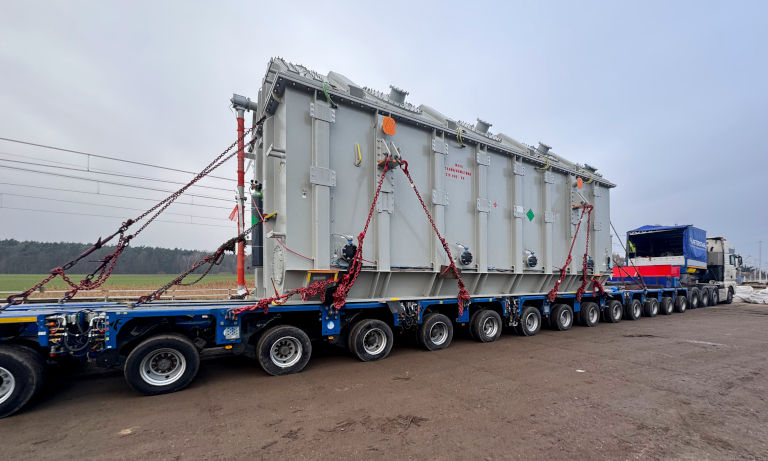Detale naczepy modułowej Faymonville MODULE MAX z załadowanym transformatorem. Widoczne liczne linie osiowe naczepy typu wózek oraz system mocowania ładunku za pomocą łańcuchów odciągowych.