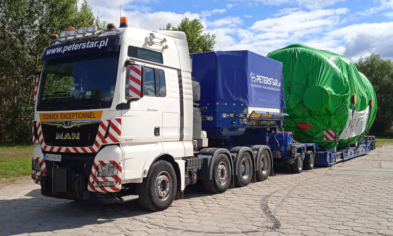 Widok z przodu na ciągnik MAN Peterstar z naczepą MODULE MAX w konfiguracji Kesselbrücke (pokład kotłowy). Transport ładunku o dużej średnicy zabezpieczonego zieloną folią ochronną, przygotowany do przejazdu nienormatywnego.